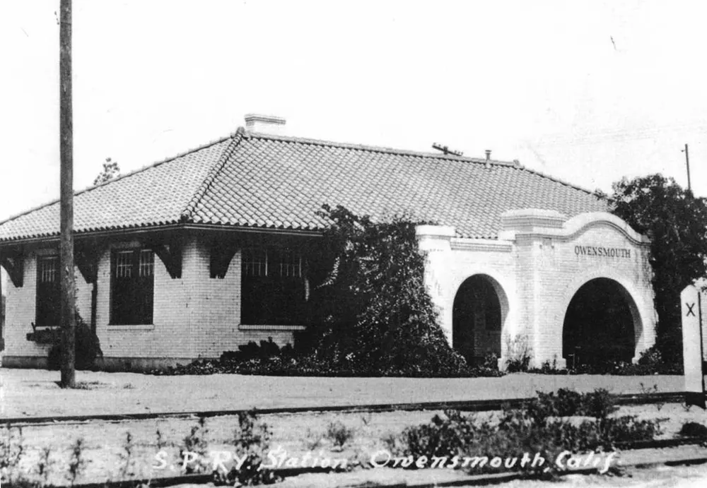 Black-and-white photo of a small tile-roofed railroad station with arched entries, about 1915